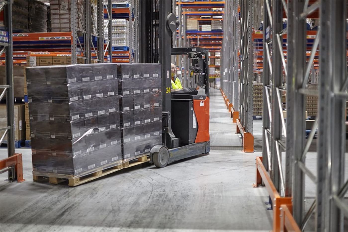 A forklift moving shrink-wrapped pallets along a racked warehouse aisle